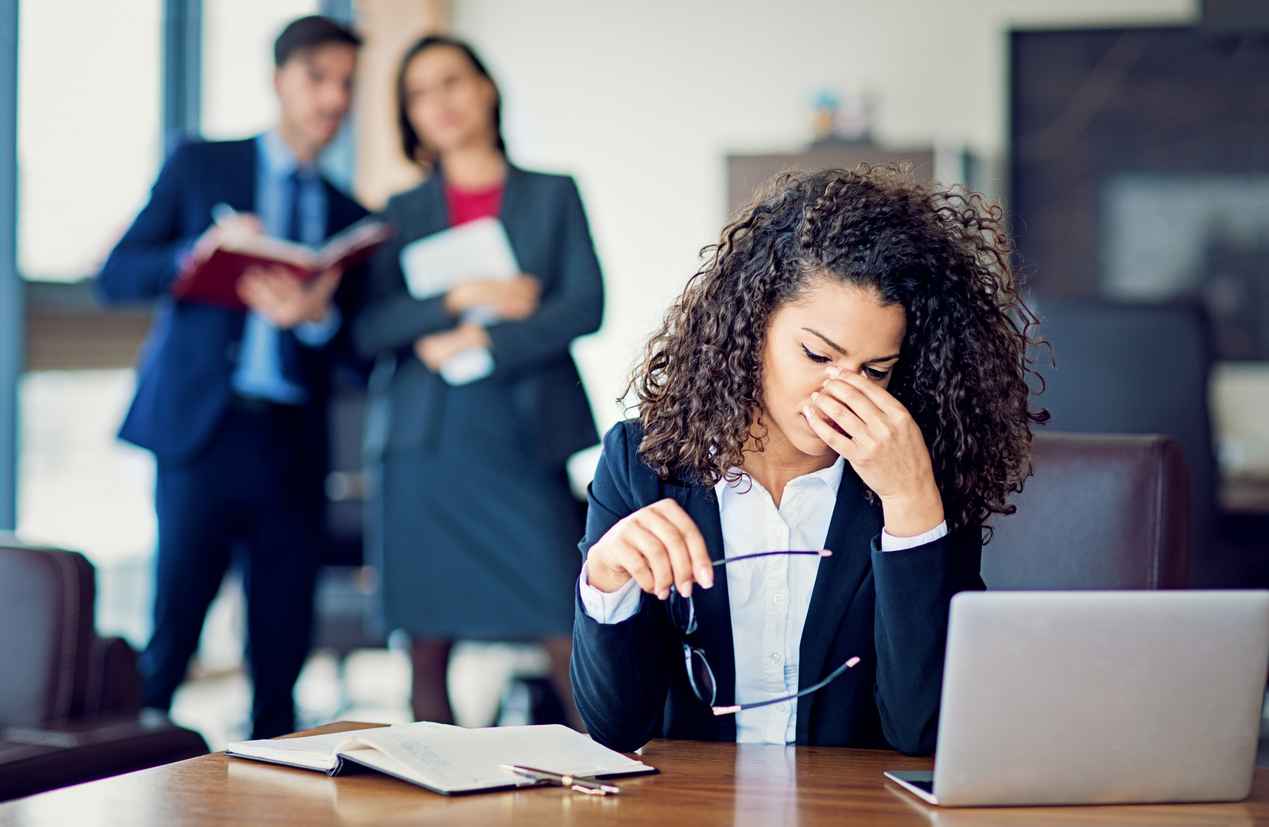 Burnout businesswoman under pressure in the office with her coworkers talking about her in the background; Handling Workplace Harassment